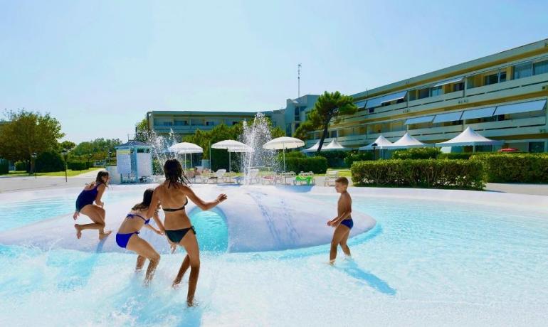 Bambini giocano in piscina con fontana, sotto il sole.